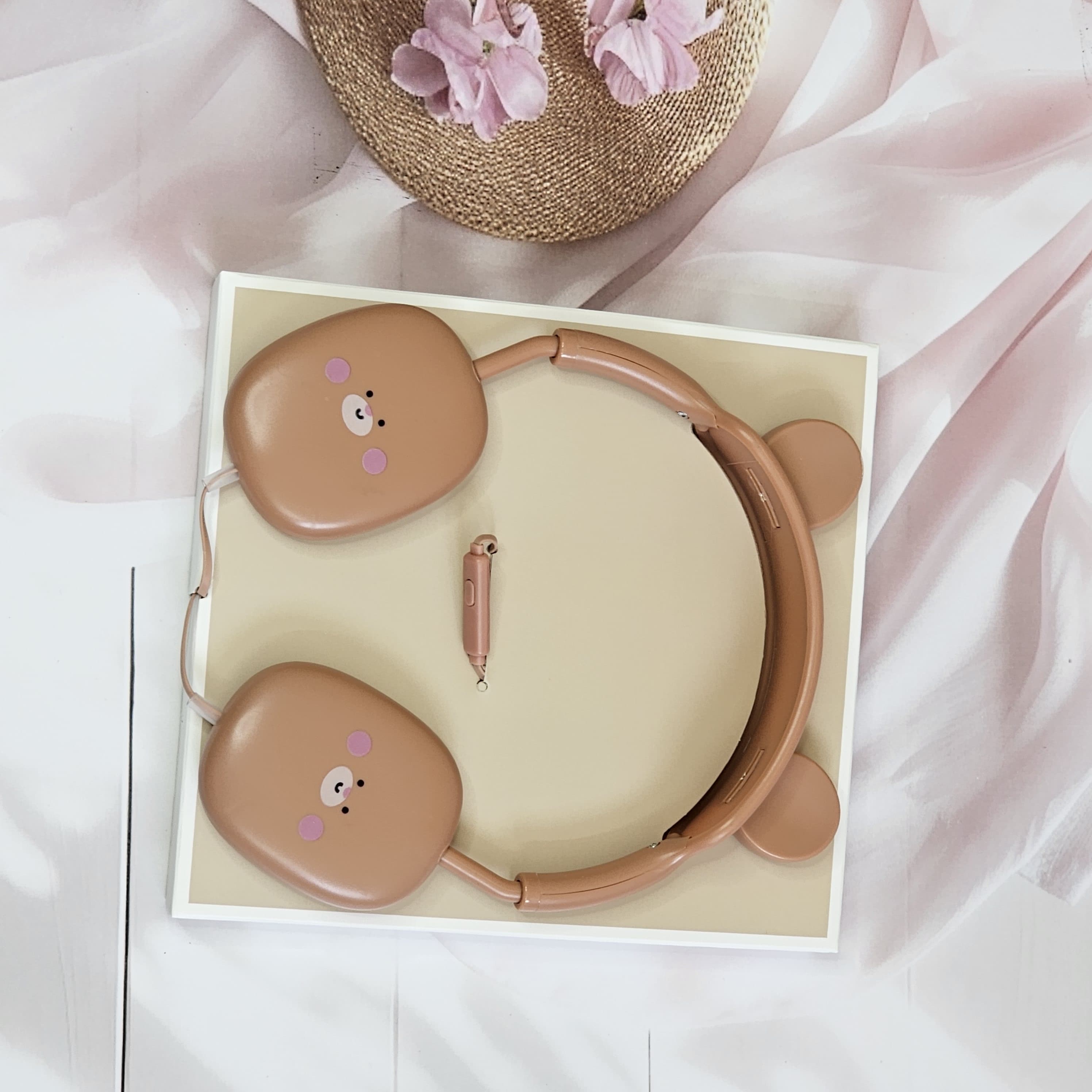 Close-up of pink BuBu Bear wired headphones for kids, showcasing the rabbit-inspired design with bear face on the earcups, a detachable microphone, and the headphones positioned on a white surface with floral accents. www.usmile.toys